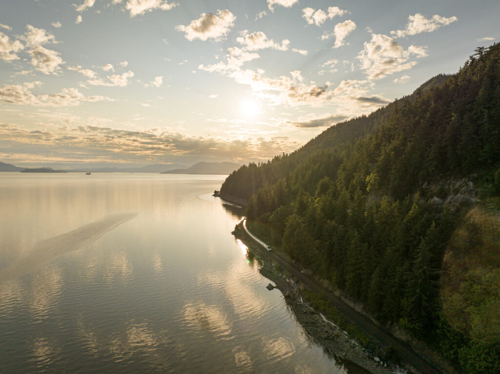 An Amtrak Cascades train glides along the edge of Puget Sound in Washington state near sunset. The pristine water is on the left, San Juan Islands are in the distance and Douglas Fir trees cover the hillside on the right. The sky is blue with thin clouds and golden sunlight filtering through. 